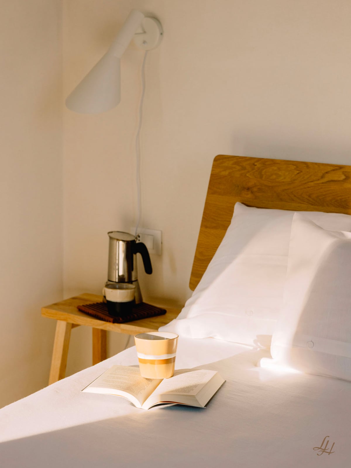 Ein Bett mit atmungsaktivem Kissenbezug Leinen, weißer Bettwäsche, einem Buch und einer Tasse sitzt im Sonnenlicht; eine Mokkakanne ruht auf dem Nachttisch.