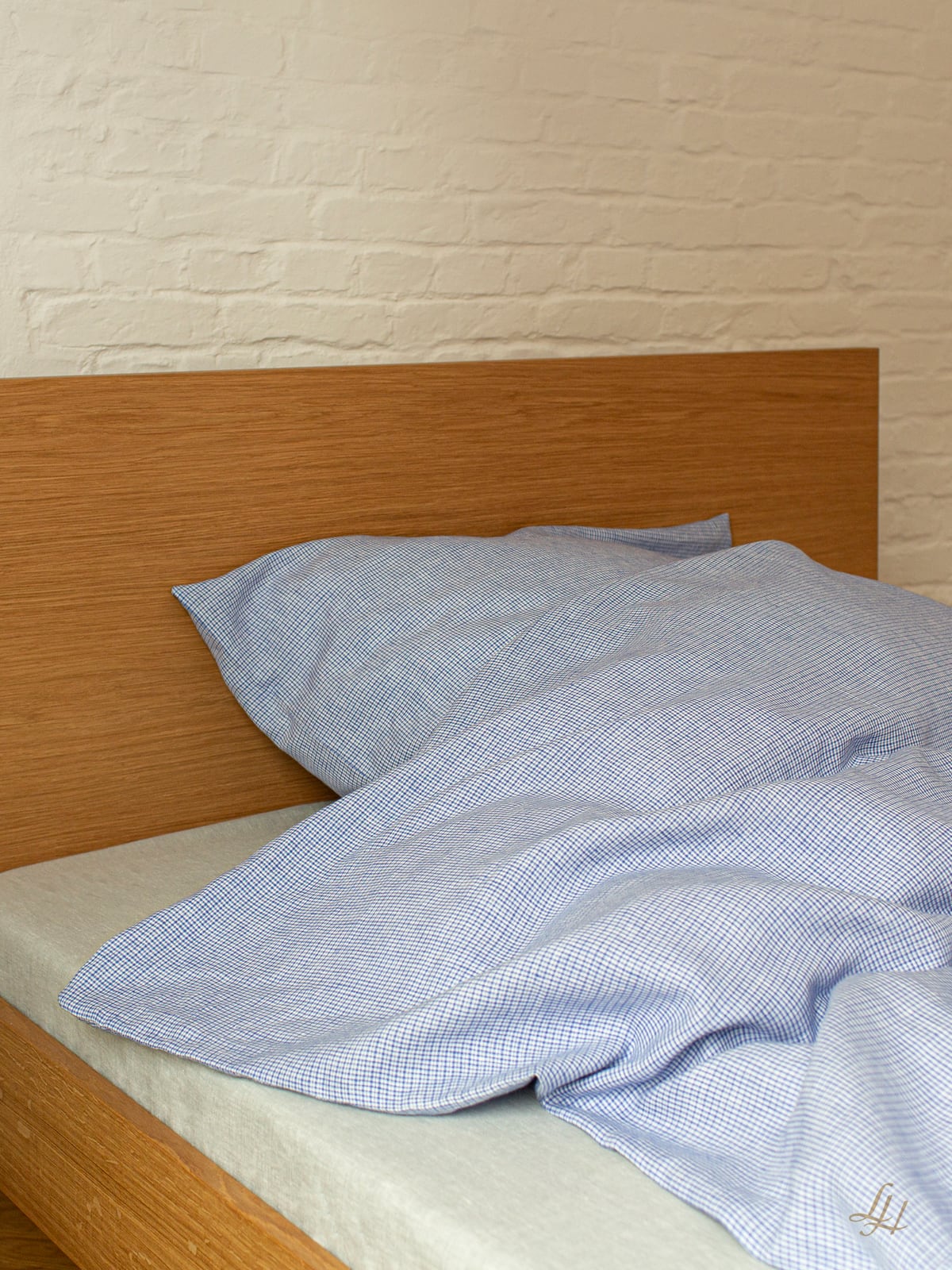 Ein ordentlich gemachtes Bett mit blau-kariertem Bettzeug und einem hölzernen Kopfteil vor einer weißen Backsteinwand.
