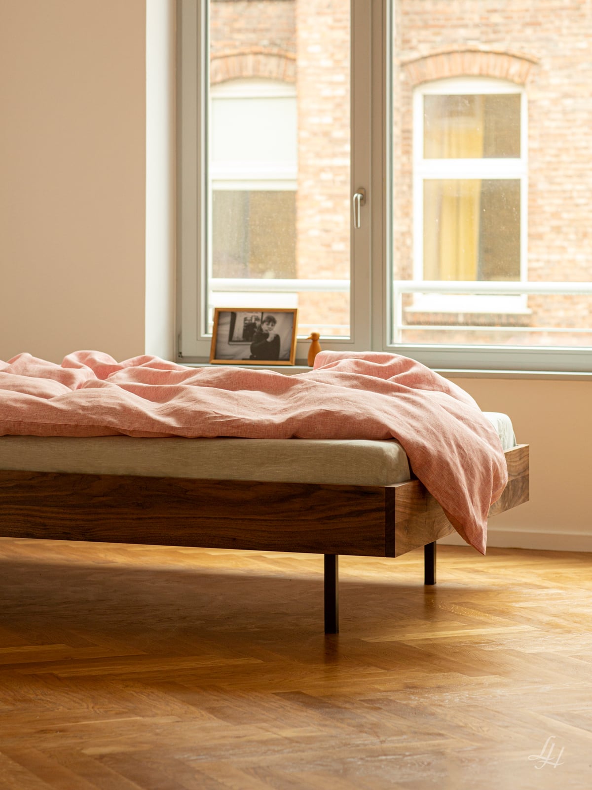 Ein Holzbett mit rosafarbenem Leinenbettbezug steht an einem Fenster, ein gerahmtes Foto auf der Fensterbank, darunter ein Holzboden. Foto von: © Sebastian Fengler
