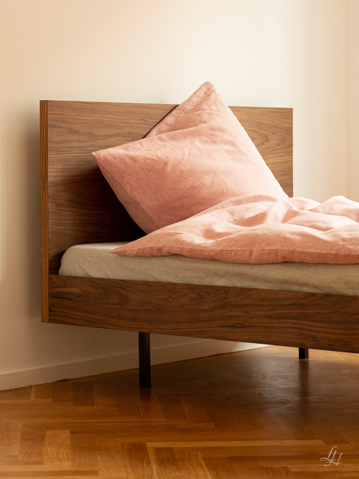 Ein Holzbett mit hohem Kopfteil, rosa Leinenbettbezug und Kopfkissen, auf Hartholzboden vor einer schlichten Wand.