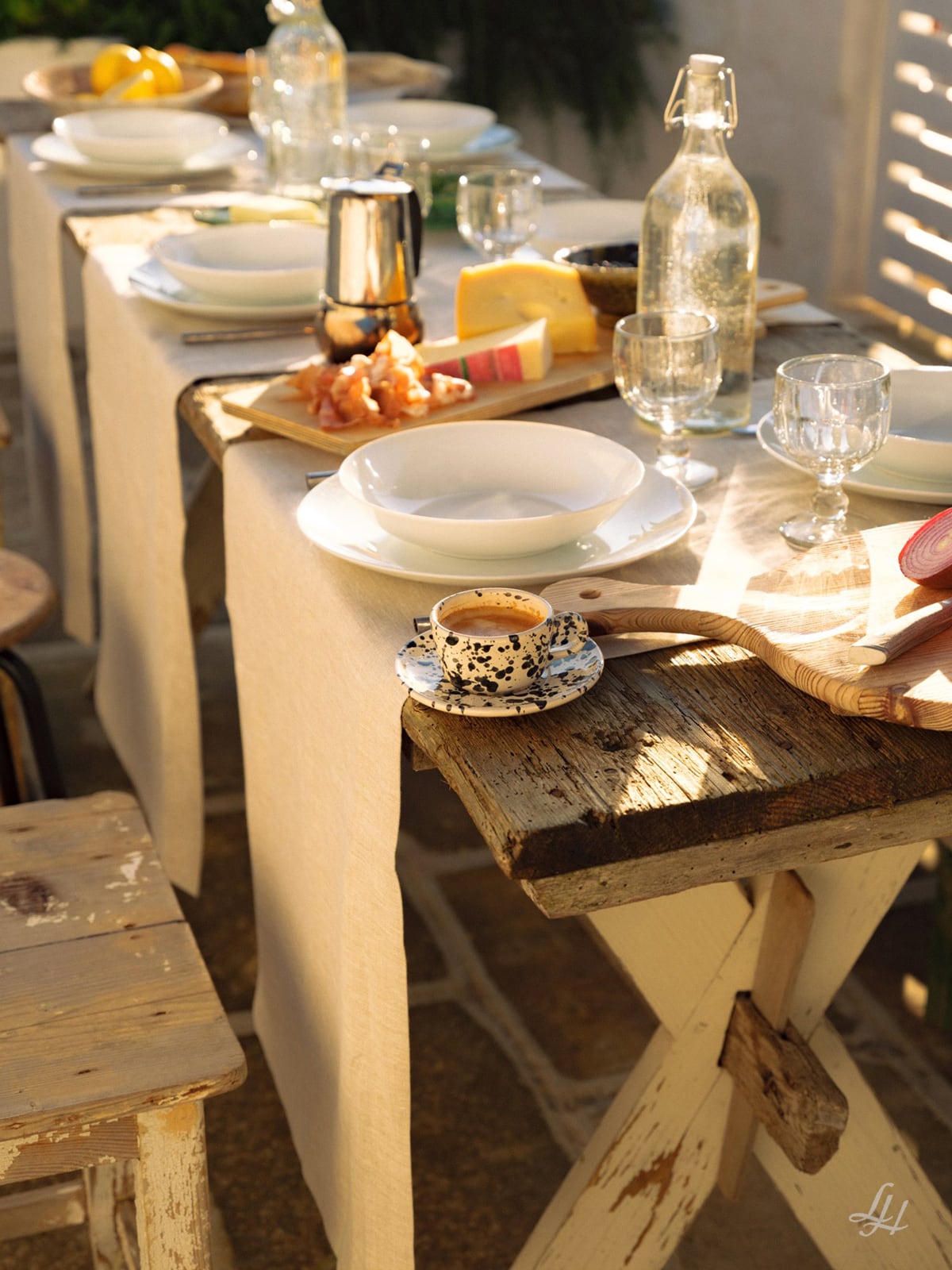Ein rustikaler Holztisch, gedeckt für eine Mahlzeit mit Tellern, Brot, Käse, Espresso und europäischer Leinen-Tischwäsche im Sonnenlicht.